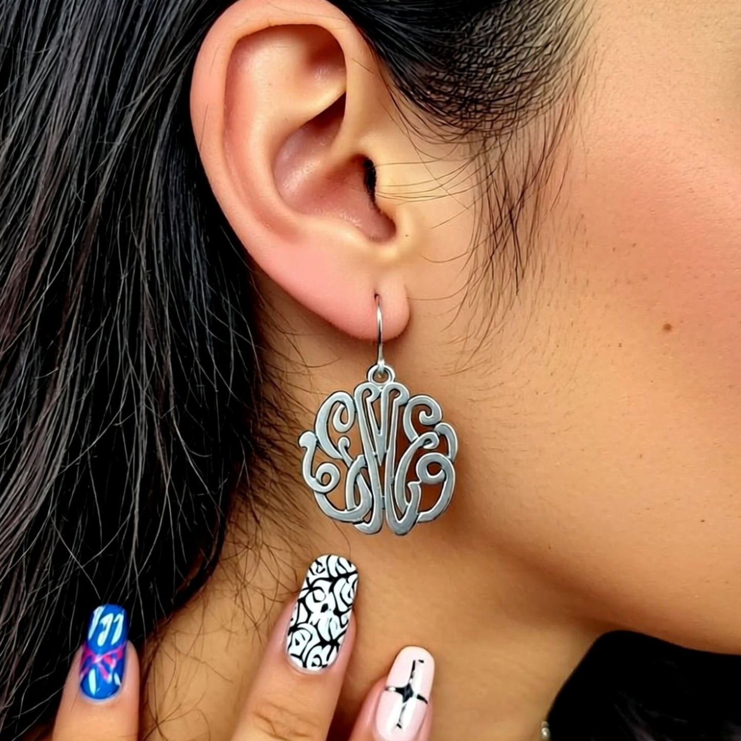 Silver monogram earrings worn by a person with black hair and multicolored nails.