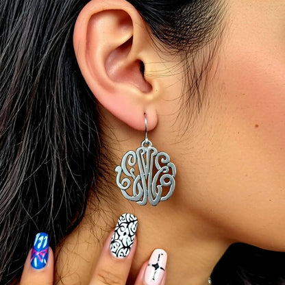 Silver monogram earrings worn by a person with black hair and multicolored nails.